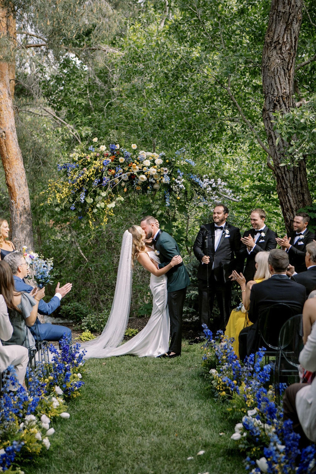 Newlywed couple shares a kiss on their wedding day in front of a stunning floral arch. Kaushay & Co Events pulls off an incredible outdoor wedding in Northern Utah. #Kaushay&Co #Kaushay&CoWeddings #Wedding #MoodySoiree #UtahWeddings #weddingplannerutah #NorthernUtahEventPlanner